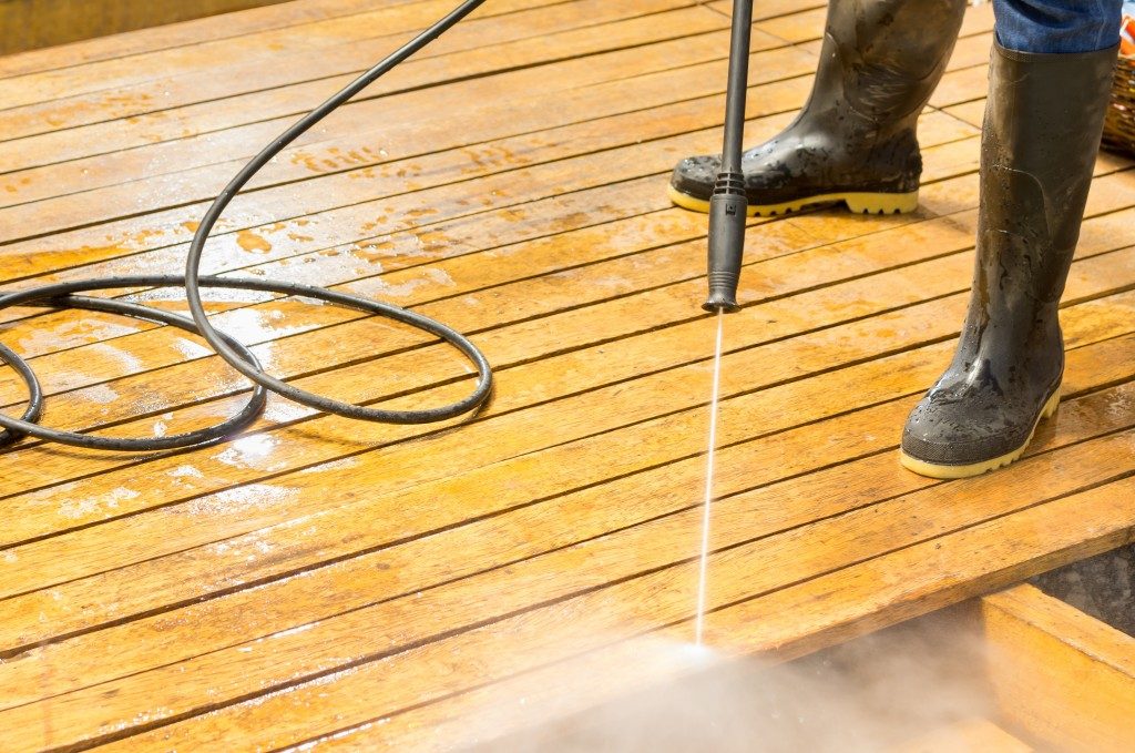 Man wearing rubber boots using high water pressure cleaner on wooden deck surface.