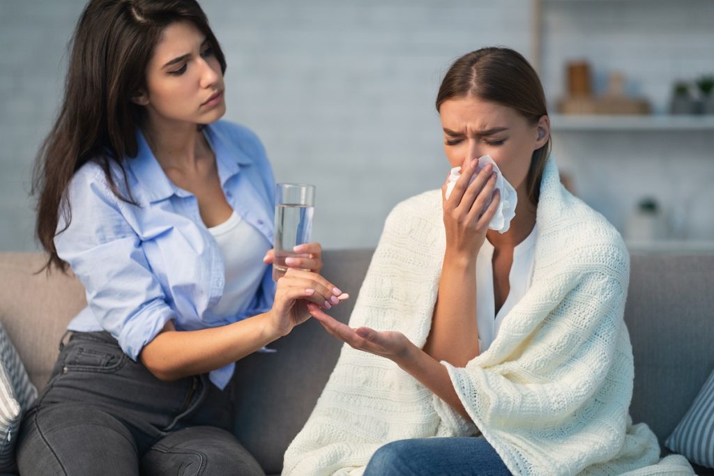 Girl Giving Pills To Sick Roommate Sitting On Couch Indoor