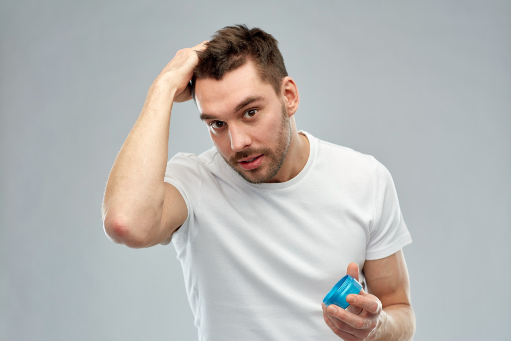 Happy young man styling his hair with wax or gel over gray background